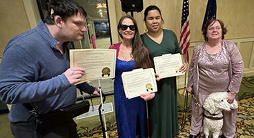 Three NFB of PA members smile and hold up certificates. Lynn stands and smiles beside them.