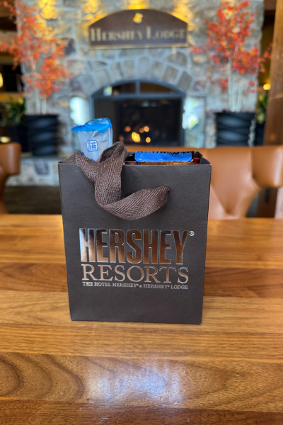 A Hershey Resorts gift bag full of chocolate candies sits on a wooden table in the Hershey Lodge. There is a stone fireplace in the background.